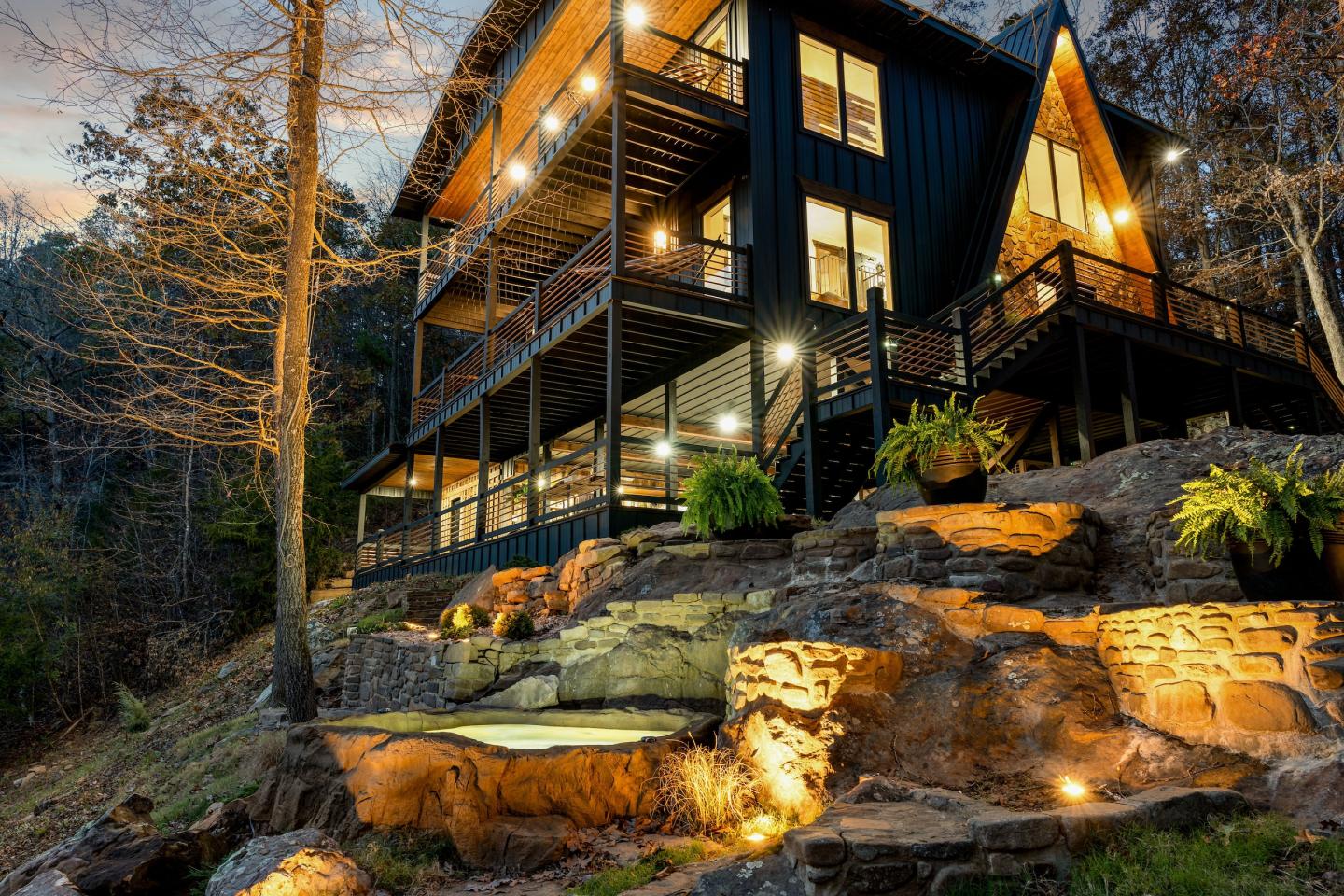 Modern black cabin with glowing windows nestled among rocky forest trees.