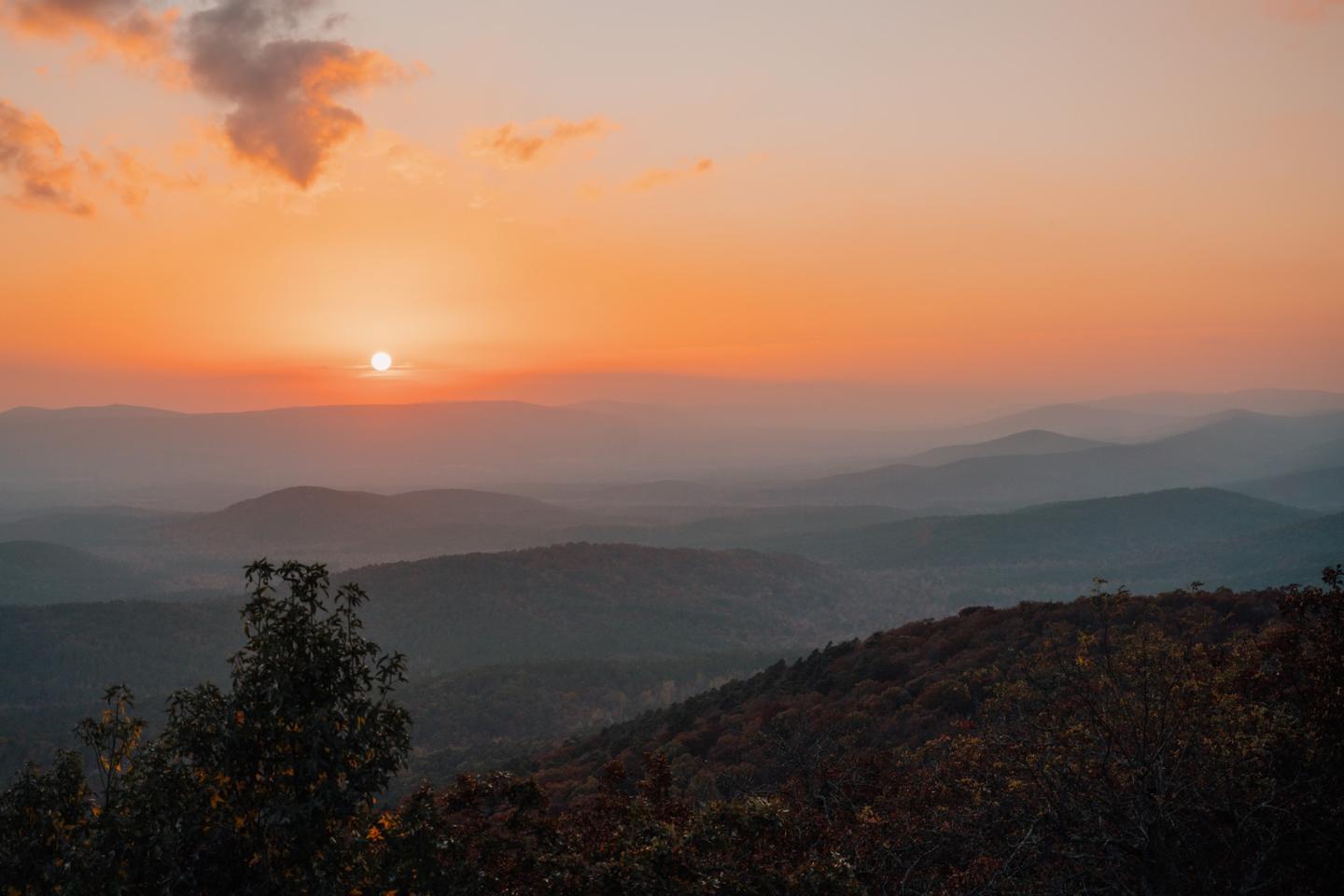 Sunset over layered mountain ridges, with silhouetted trees in the foreground.