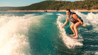 A lake setting near Mountain Home, Arkansas. Blue sky and green-tree hills are seen in the background. Blue water and white wake fill the rest of the photograph. A girl is seen on a board surfing in the wake. Photo source: Magnetic Creative.