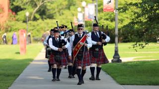 A traditional Scottish bagpiping band is leading a procession of graduates down the sidewalk on the Lyon College campus. All wearing kilts, the man in front is wearing a sash and carrying a baton. Three pipers are walking behind and playing, as well as a drummer. Street lamps are adorned with red and blue banners.